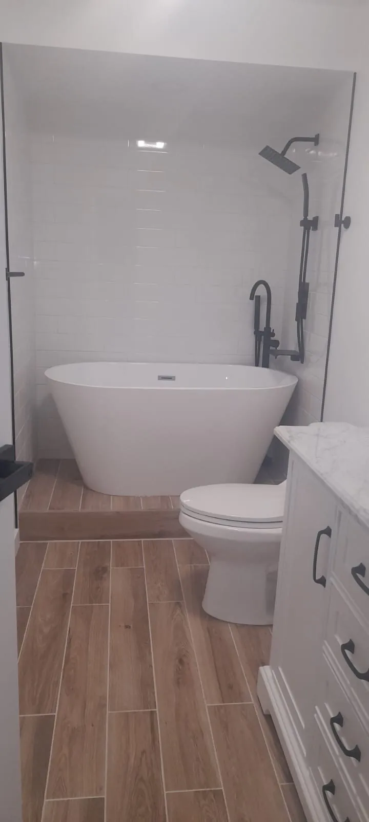 A bathroom with a white freestanding tub, a black shower set and wood-look floor tile