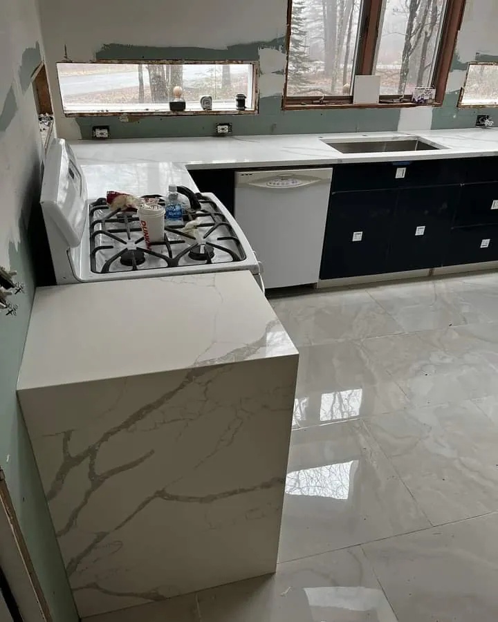A kitchen during the finish: a white quartz island with a waterfall edge, a gas range, black lower cabinets and large marble-look floor tile